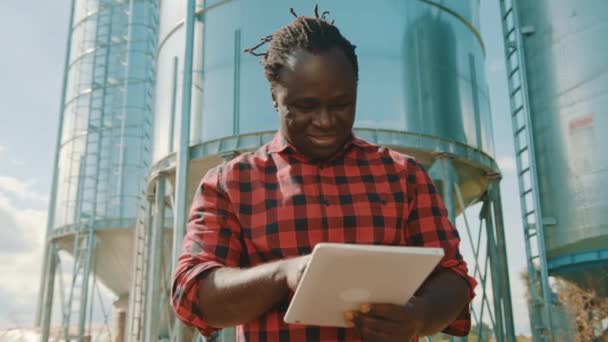 Genç, yakışıklı Afrikalı bir adam. Silo sisteminin önünde tablet kullanan bir mühendis. — Stok video