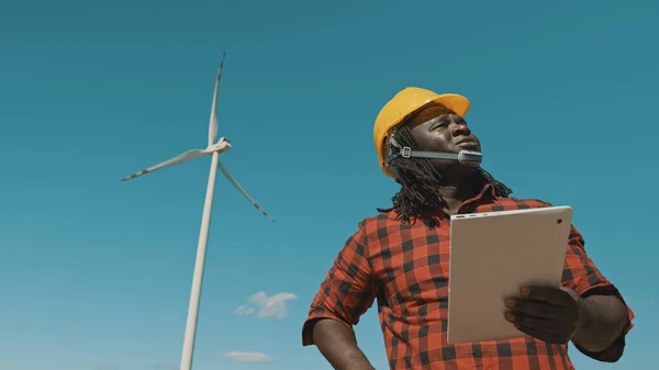 Felnőtt vagy érett professzionális afrikai férfi kemény kalapban áll modern digitális táblagéppel ellen szélmalom erőmű ellenőrző tárgyak — Stock Fotó
