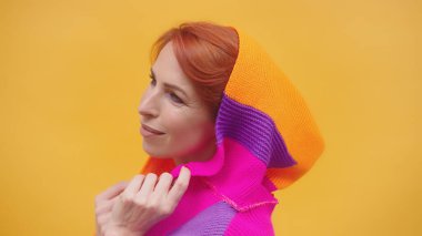 Sarı arka planda, renkli kapüşonlu, orta yaşlı, şık, kızıl saçlı bir kadın.