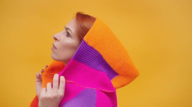 Sarı arka planda, renkli kapüşonlu, orta yaşlı, şık, kızıl saçlı bir kadın.