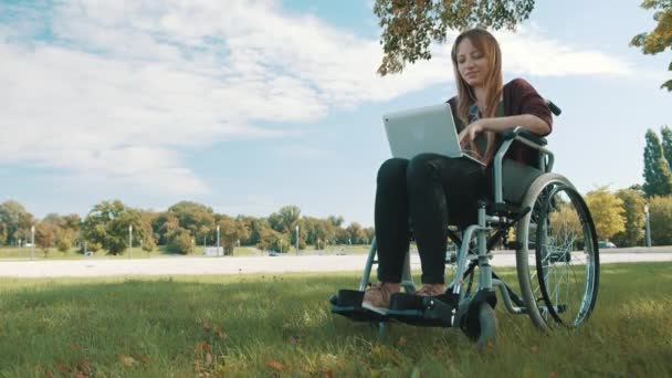 Concepto de trabajo remoto. Joven hermosa mujer caucásica discapacitada en silla de ruedas utilizando el ordenador portátil en la naturaleza — Vídeo de stock