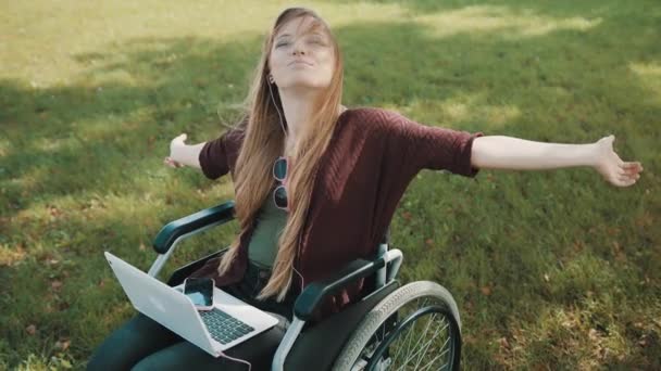 Joven mujer caucásica discapacitada en silla de ruedas con portátil disfrutando del día de otoño en el parque con música desde el teléfono inteligente — Vídeo de stock