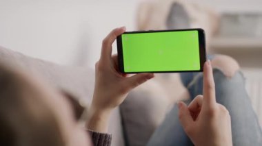 Kanepede dinlenirken ve el sallarken elinde akıllı telefon yeşil ekranı tutan genç bir kadın. Video çağrısı