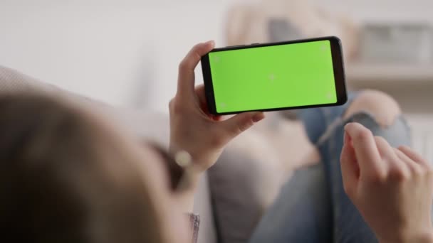 Jeune femme tenant smartphone écran vert tout en se reposant sur le canapé. Regarder des films au téléphone — Video