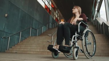 Merdiven önünde tekerlekli sandalyede oturan, cesareti kırılmış, beyaz bir kadın.