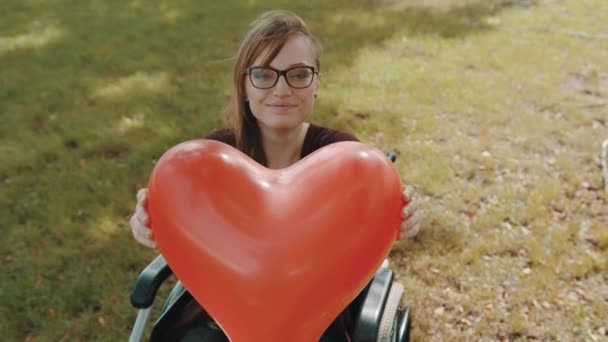 Beautiful young woman in the wheelchair holding heart shaped balloon in the park — Stock Video