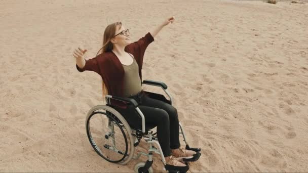 Joven mujer discapacitada en la playa de arena sentada en la silla de ruedas disfrutando del soleado día de otoño — Vídeos de Stock