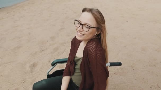 Genç engelli kadın kumlu sahilde oturuyor tekerlekli sandalyede güneşli sonbahar gününün tadını çıkarıyor.. — Stok video