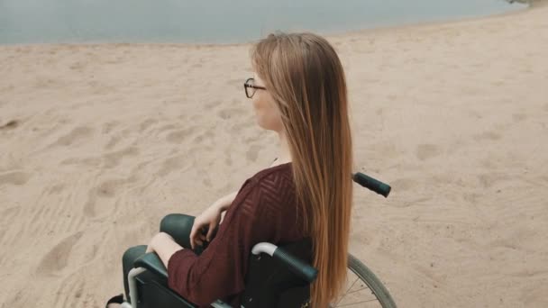 Młoda, szczęśliwa, biała kobieta na wózku inwalidzkim na piaszczystej plaży, robiąca serce rękami, ujęcie z ramienia — Wideo stockowe