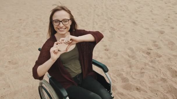 Steun voor gehandicapten. Jonge vrouw in de rolstoel aan de zandige rivierkust toont hart met handen — Stockvideo