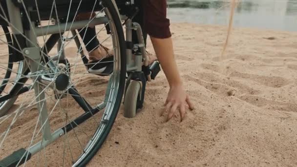 Primer plano, mano de minusválidos en silla de ruedas jugando con la arena en la costa del río. Vacaciones y viajes con discapacidad — Vídeo de stock