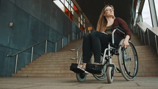 Young discouraged caucasian woman in the wheelchair in front of the staircase looking around for accessible way — Stock Photo, Image