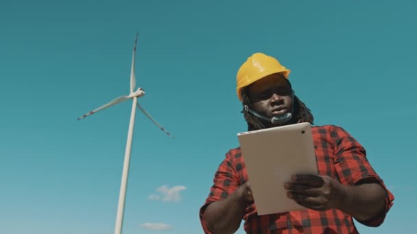 Afrikansk man i hård hatt stående med modern digital tablett mot väderkvarn kraftverk gör inspektioner av objekt — Stockvideo