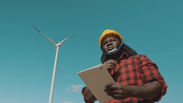 Profesionální africký muž v tvrdé čepici stojící s moderním digitálním tabletem proti větrné elektrárně dělat kontroly objektů — Stock video