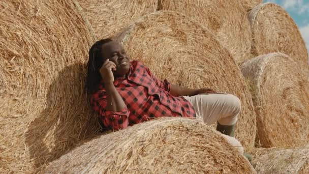 Jovem agricultor africano feliz deitado na pilha de rolos de feno, descansando e falando ao telefone — Vídeo de Stock