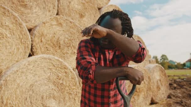 Jeune agriculteur africain nettoyer la sueur avec la main de son front tout en se reposant sur la poignée de fourche. Foin rouleau pile en arrière-plan — Video