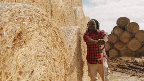 Joven agricultor africano descansando en la manija de la horquilla frente a la pila de rollo de heno — Vídeos de Stock