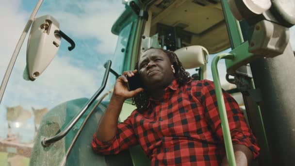 Ung afrikansk bonde talar på smartphone medan lutar sig mot den gröna traktorn — Stockvideo