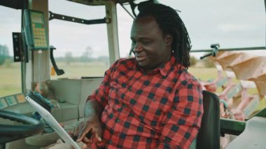 Genç Afrikalı çiftçi traktör kabininde otururken tablet kullanıyor ve gülümsüyor.