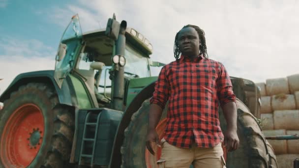 Africano, fazendeiro em pé na frente do trator verde e cruzando as mãos. Fay carretéis pilha no fundo — Vídeo de Stock
