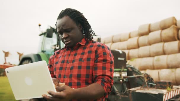 Fiatal afrikai mezőgazdasági termelő használ tabletta előtt og nagy zöld traktor anf szénakazal — Stock videók