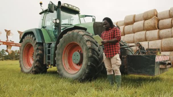 Młody rolnik afrykański za pomocą tabletki w przedniej og duży zielony ciągnik i stóg siana — Wideo stockowe