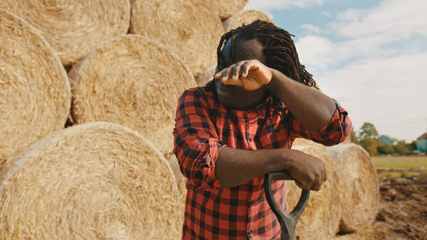 Un joven agricultor africano limpiando el sudor con la mano de la frente. Pila de rollo de heno en el fondo — Foto de Stock