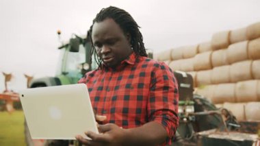 Genç Afrikalı çiftçi büyük yeşil traktör ve samanlığın önünde tablet kullanıyor.