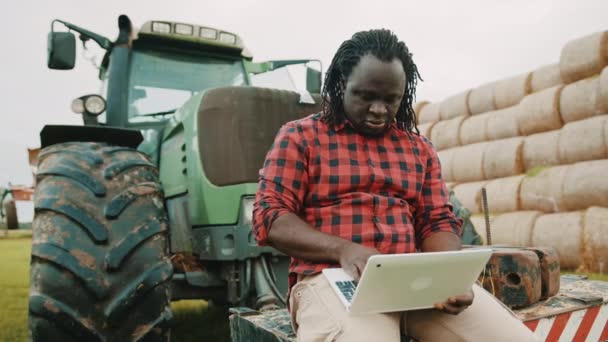Young african farmer using tablet while sitting on green tractor. Haystack in the background — Stock Video