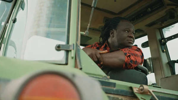 Cansado jovem fazendeiro africano sentado na cabine do trator e olhando ao redor — Fotografia de Stock