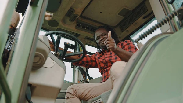 Joven agricultor africano sentado en la cabaña del tractor y mostrando los pulgares hacia arriba como aprobación — Foto de Stock
