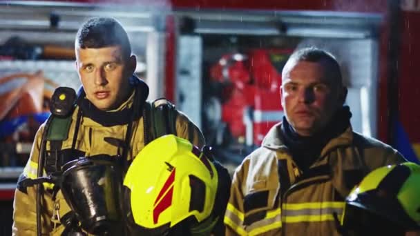 Ritratto di due giovani vigili del fuoco sotto la pioggia davanti al motore dei pompieri in uniforme. Trapano antincendio — Video Stock