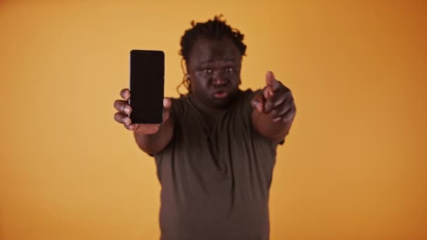 Hombre africano enojado sosteniendo el teléfono inteligente y señalando los dedos hacia la cámara — Vídeos de Stock