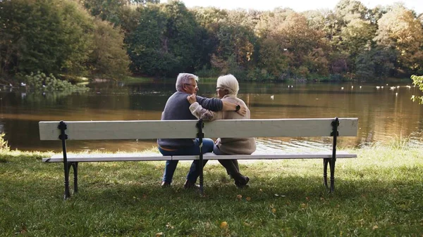 Yaşlı gri saçlı çift sonbahar günü nehir kenarındaki bankta oturuyor. Romantizm ve yaşlılık — Stok fotoğraf