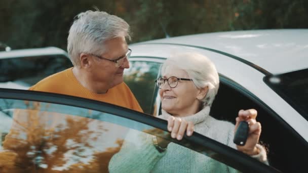 Una pareja de ancianos compró un auto nuevo. De pie cerca de la puerta y la mujer saludando con llaves — Vídeos de Stock