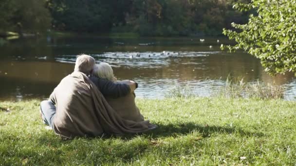 Пожилая серая пара, сидящая на скамейке у реки в осенний день, покрытая одеялом. Романтика в старости — стоковое видео