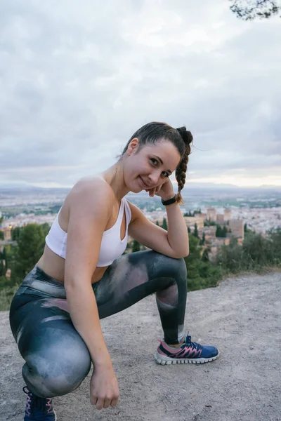 Young athlete girl crouched posing with the city of Granada and the Alhambra in the background in the afternoon in summer. Young woman smiling and enjoying in nature.