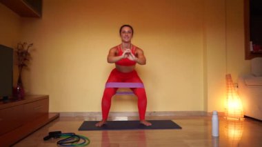 Çömelme hareketleri yapan genç bir kadın. Evdeki oturma odasındaki paspasın üzerinde spor giyimi eğitimi alan zinde bir kız.