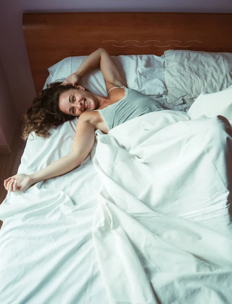 Portrait of pretty young girl looking at camera and laughing in morning and lying on white bed duvet.