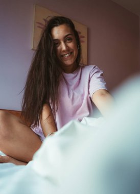 Pijamalı, gülümseyen genç bir kadın, yatak odasında beyaz çarşaflarla, yatağında oturuyor..
