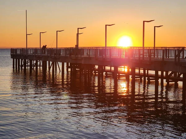 a promenade pier jutting out into the sea against the backdrop of a bright orange sunset