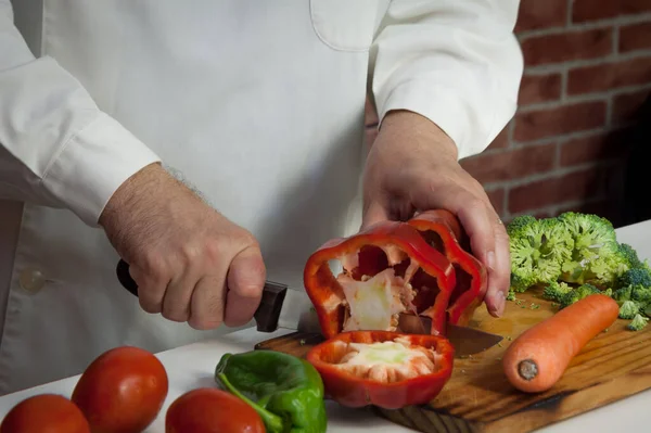 man cooking and cutting vegetables, cauliflower, bell pepper, carrot, green pepper and tomatoes, on wooden board and brick background, white shirt, horizontal format