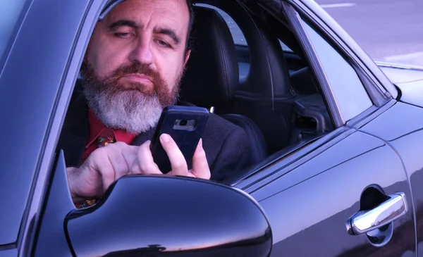stylish executive man with beard inside black sports car with phone and typing message on whatsapp