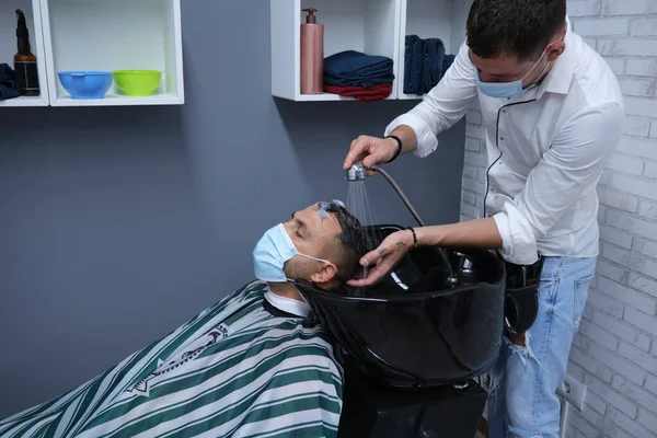 Young barber with face mask washing hair carefully young boy with face mask, horizontal format