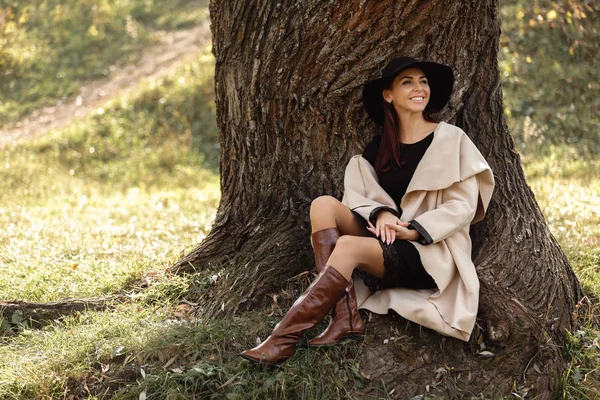 Woman in a beige coat and black hat sitting under tree — Stock Photo, Image