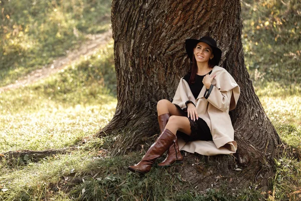 Frau in beigem Mantel und schwarzem Hut sitzt unter Baum — Stockfoto