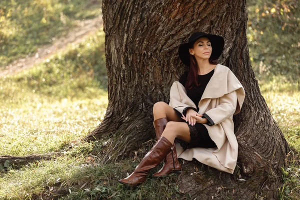 Frau in beigem Mantel und schwarzem Hut sitzt unter Baum — Stockfoto