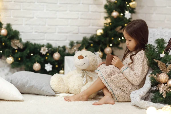 Cute girl drinks hot cocoa and plays with teddy bear at home. Christmas — Stock Photo, Image