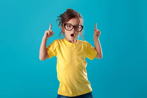 Mignonne surprise petite fille en lunettes et t-shirt jaune pointant vers le haut — Photo