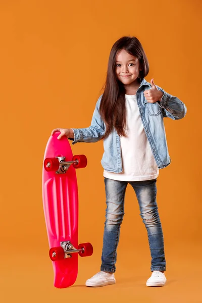 Стильна маленька дівчинка з скейтбордом в денімі на помаранчевому фоні — стокове фото